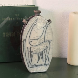 以下が含まれることがあります： クリーム色の表面と黒い線画の人物が描かれた、楕円形のセラミック花瓶。花瓶は濃い茶色の縁と上部に小さなハンドルが付いています。白いろうそくと緑の本が背景にあります。