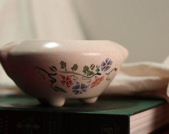 May include: A white ceramic bowl with a floral design featuring pink, blue, and green flowers. The bowl is sitting on a green book.
