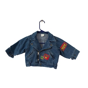 May include: A blue denim jacket with a zipper closure and two front pockets. The jacket features colorful patches with text and designs, including a heart with wings and a star. It is hanging on a blue hanger.