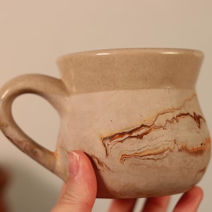 Pode incluir: Uma caneca de cerâmica bege com uma alça curva. A caneca apresenta um padrão marmorizado em tons de castanho e bege. A borda superior é bege claro sólido. A caneca é segurada por uma mão.