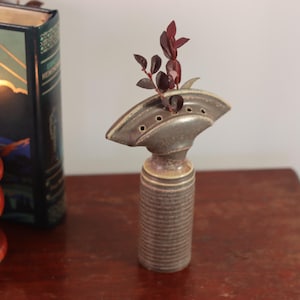 May include: A brown ceramic vase with a unique design, featuring a wide, flat top with holes and a cylindrical base. The vase is filled with a small arrangement of burgundy-colored leaves.