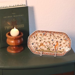 May include: An octagonal ceramic serving platter with a detailed floral and animal design. The platter features a brown border and a cream-colored base with illustrations of birds, deer, and flowers. A wooden candle holder and a book are in the background.