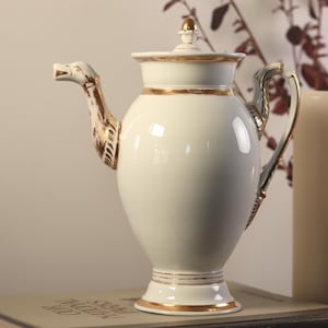 Puede incluir: Una tetera de porcelana blanca con detalles dorados y un pico decorativo. La tetera tiene un cuerpo redondo y un pico alto y delgado. La tetera está sobre un libro.