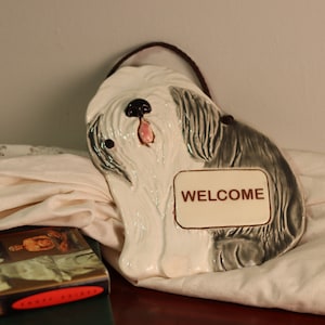 May include: Ceramic wall hanging of a white and gray Old English Sheepdog with a welcome sign. The dog is lying down with its head turned to the side and its tongue sticking out.