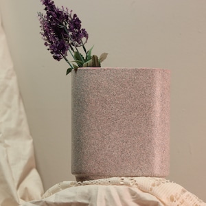 May include: A pink speckled ceramic vase with a square shape, holding a bouquet of purple flowers.