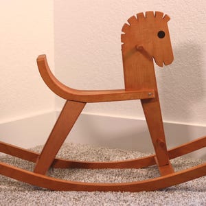 Könnte beinhalten: Ein hellbraunes Schaukelpferd aus Holz mit einem stilisierten Kopf, einem dunklen kreisförmigen Auge, einer gekerbten Mähne, einem geschwungenen Sitz und zwei langen, gebogenen Kufen. Die Struktur ist mit sichtbaren Metallbeschlägen verbunden.