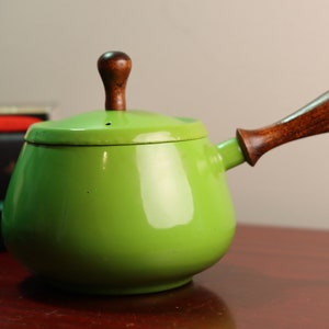 Puede incluir: Una cacerola de esmalte verde brillante con un mango y una tapa de madera. La cacerola tiene un cuerpo redondeado y un pequeño pico.