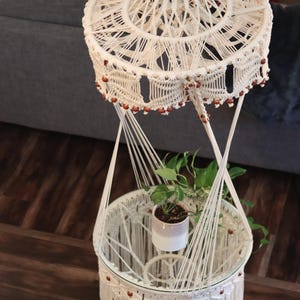 May include: A cream-colored macrame plant stand with a glass tabletop and a hanging design. The stand features intricate knotwork, wooden beads, and a small potted plant. The overall design is bohemian and decorative.