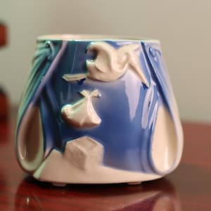 Peut inclure: Un pot en céramique bleu et blanc avec un motif décoratif d'oiseaux et de fleurs stylisés. Le pot a une forme arrondie et une finition lisse et brillante.