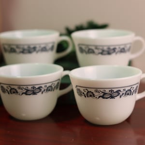 Puede incluir: Cuatro tazas de té de cerámica blanca con diseños florales azules y asas. Las tazas están dispuestas sobre una superficie de madera.