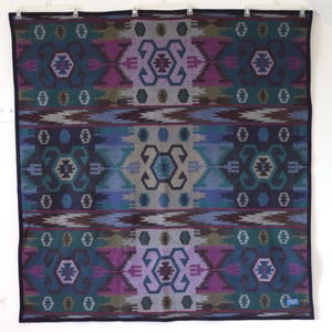 May include: A rectangular woven blanket featuring a repeating geometric pattern in shades of teal, purple, blue, and grey. The design includes symmetrical hexagonal and angular motifs arranged in horizontal bands with a dark blue border edge.