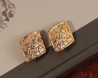 May include: Two gold-coloured square earrings with a textured surface and embossed design. The earrings have a vintage aesthetic and are displayed on a neutral background.