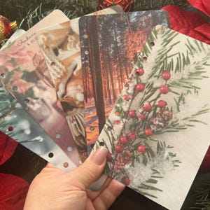 May include: Five different planner pages with various designs. The pages are white with a hole punch on the left side. The designs include a forest scene, a snowy pine branch, a floral design, and a design with a woman's face. The text on one of the pages reads "Anders Home & Hue".