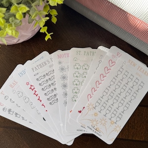 May include: A selection of white planner stickers with diverse designs and text, including holiday themes such as Halloween, Father's Day, and St. Patrick's Day. The stickers feature illustrations of ghosts, stars, clovers, and champagne glasses.
