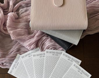 May include: A light pink wallet with a textured surface and a snap closure, stacked on top of two other wallets. Below are several white cards with savings challenge designs, each with a different amount to save.
