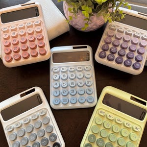 Puede incluir: Cinco calculadoras de colores pastel con marcos blancos y botones de varios colores están dispuestas sobre una superficie marrón oscuro. Las calculadoras tienen una pantalla digital y teclas de función estándar. Una pequeña planta en una maceta rosa está en el fondo.