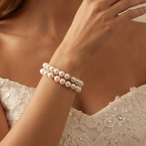 May include: Two pearl bracelets with gold details are worn on a wrist. The bracelets are made of round, white pearls. The background shows a white, lace-detailed dress.