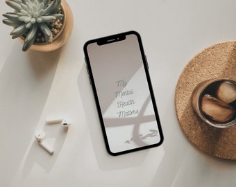 以下が含まれることがあります： 白い画面に「My Mental Health Matters」という文字が表示された黒いスマートフォン。スマートフォンは、多肉植物と氷水が入ったグラスを置いたコースターのある白い表面の上に置かれている。
