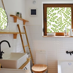 May include: A white bathroom with a white toilet, a white bathtub, a wooden ladder, a wooden shelf with a round mirror, and a window with a green leafy pattern.