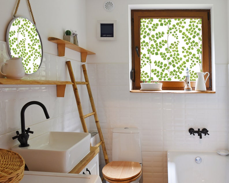 May include: A white bathroom with a white toilet, a white bathtub, a wooden ladder, a wooden shelf with a round mirror, and a window with a green leafy pattern.