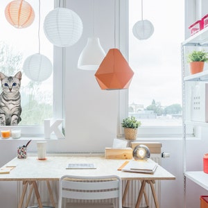 May include: A white desk with a light wood top and a white chair. There is a cat looking out the window, a lamp, and a shelf with various items on it.
