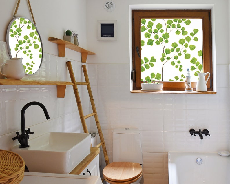 May include: A white bathroom with a white toilet, a white bathtub, a white sink, a wooden ladder, a round mirror with a leafy design, and a window with a leafy design.