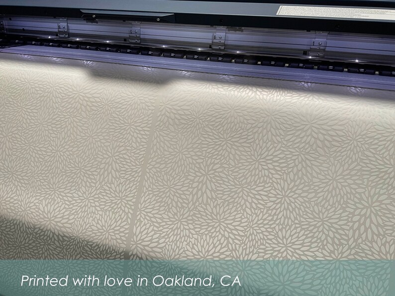May include: A large format printer printing a white fabric with a delicate floral pattern. The fabric is being printed with a white ink on a light beige background. The text "Printed with love in Oakland, CA" is visible at the bottom of the image.