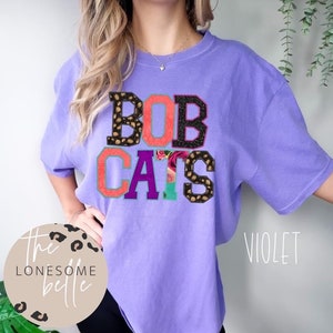 May include: A purple t-shirt with the words "BOB CATS" in colorful, patterned letters. The letters are layered on top of each other and have a vintage, distressed look.