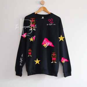 May include: Black sweatshirt with a design featuring a bear wearing sunglasses, stars, hearts, and pennants. The text "go fight win" and "Bears" are also included in the design. The sweatshirt is hanging on a wooden hanger.