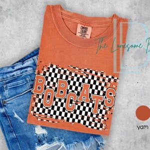 May include: A burnt orange t-shirt with a distressed checkered pattern and the text "BOBCATS" in white outlined letters. The t-shirt is folded and laid on top of a pair of blue jeans.