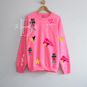 May include: Bright pink sweatshirt with various appliqu&eacute;s, including a cartoon bulldog wearing sunglasses and shorts, gold stars, black hearts, and pennants that say "Bulldogs". The words "go fight win" are also on the sweatshirt.