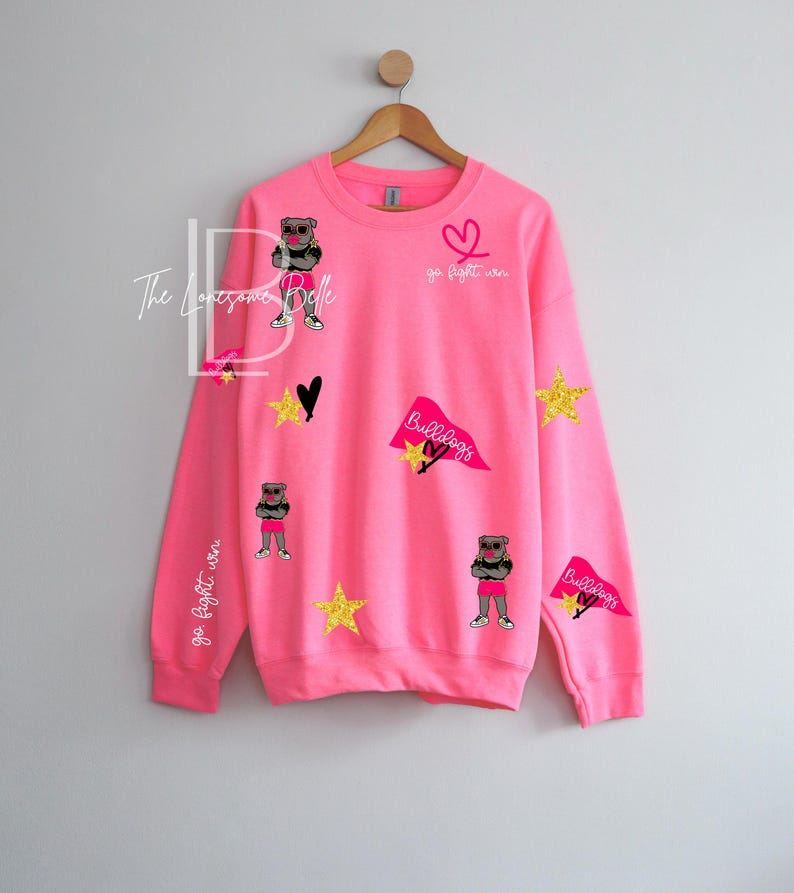 May include: Bright pink sweatshirt with various appliqu&eacute;s, including a cartoon bulldog wearing sunglasses and shorts, gold stars, black hearts, and pennants that say "Bulldogs". The words "go fight win" are also on the sweatshirt.