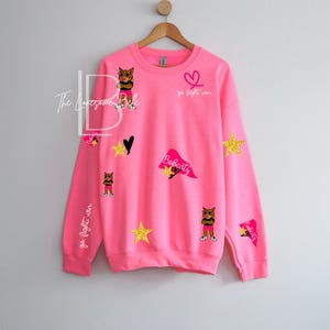May include: A bright pink sweatshirt with various appliqu&eacute;s, including gold stars, a black heart, and cat illustrations. The text "Go Fight Win" and "Bobcats" are also visible. The sweatshirt is hanging on a wooden hanger.