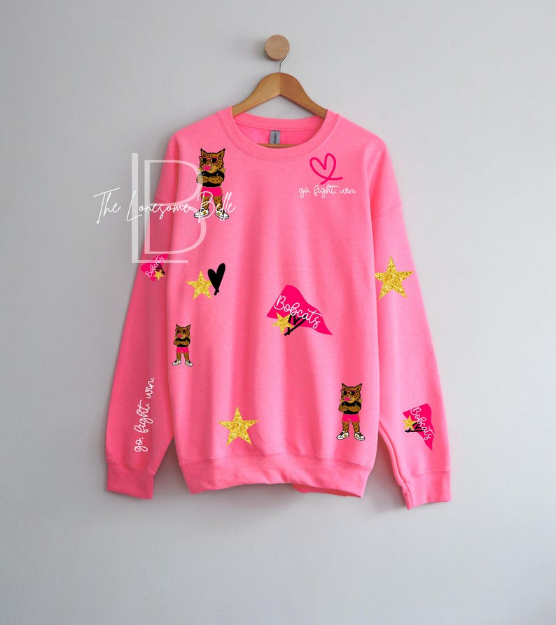 May include: A bright pink sweatshirt with various appliqu&eacute;s, including gold stars, a black heart, and cat illustrations. The text "Go Fight Win" and "Bobcats" are also visible. The sweatshirt is hanging on a wooden hanger.