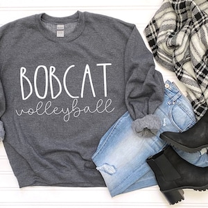May include: Gray sweatshirt with white text that reads "BOBCAT volleyball". The sweatshirt is laid out on a white surface with a pair of black ankle boots, blue jeans, and a black and white plaid scarf.