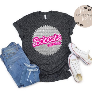 May include: A black and grey leopard print t-shirt with a silver glitter circle design that says "Bobcats only in Gary" in pink letters.