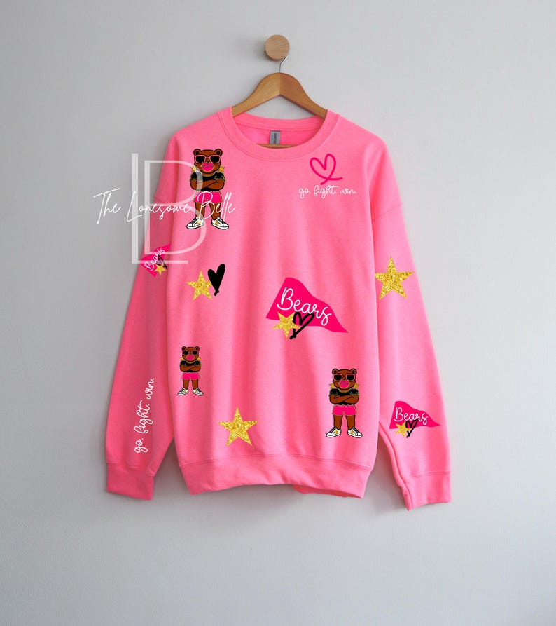 May include: A bright pink sweatshirt with various graphics. The design includes a cartoon bear wearing sunglasses, stars, hearts, and pennants that say "Bears". The text "go fight win" is also visible.