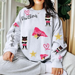 May include: A light gray sweatshirt with a playful design featuring the word "Panthers" in black script, pink hearts, gold stars, and cartoon cat illustrations wearing sunglasses. The sleeve has the text "go. fight. win."