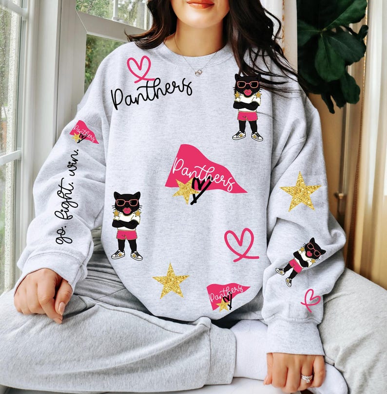 May include: A light gray sweatshirt with a playful design featuring the word "Panthers" in black script, pink hearts, gold stars, and cartoon cat illustrations wearing sunglasses. The sleeve has the text "go. fight. win."