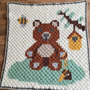 Può includere: Una copertina per neonati all'uncinetto con un orso marrone, un'ape e un alveare. La copertina è realizzata con un punto a trama e presenta una combinazione di colori verde, marrone, giallo e bianco.