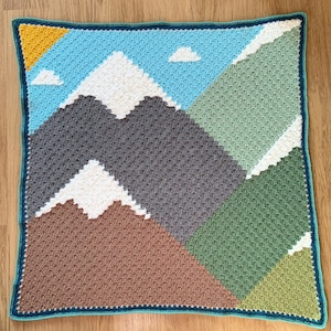 Peut inclure: Une couverture au crochet représentant une scène de montagne. La couverture présente un ciel bleu avec des nuages blancs, des montagnes brunes et grises et des collines vertes et brunes. La couverture est bordée d'un bord au crochet bleu sarcelle.