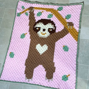 Puede incluir: Una manta de crochet rosa con un perezoso marrón colgado de una rama. El perezoso tiene un corazón blanco en el pecho.