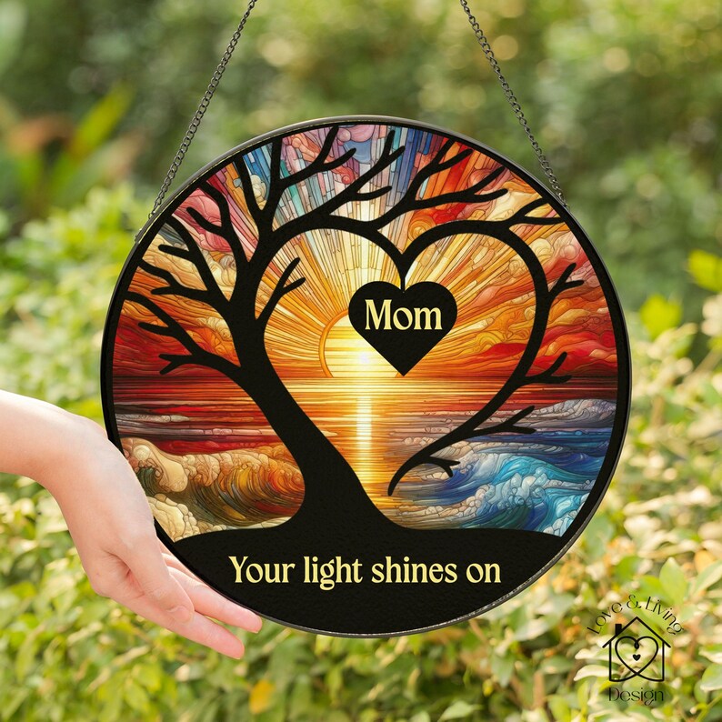 May include: Round stained glass suncatcher with a black tree design. The tree's branches form a heart shape with the word "Mom" inside. The background features a sunset over the ocean, with the text "Your light shines on".