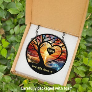 May include: A round, stained glass suncatcher with a tree design, a heart with the word "Mom", and the text "Your light shines on". The suncatcher is in a cardboard box with foam packaging, surrounded by green leaves.