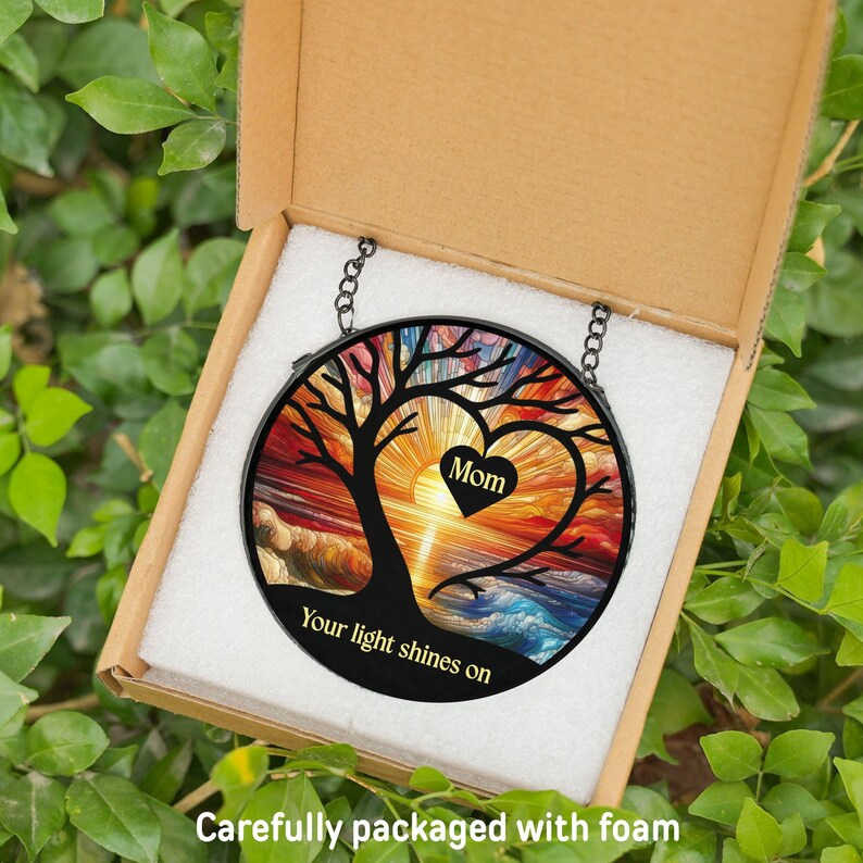 May include: A round, stained glass suncatcher with a tree design, a heart with the word "Mom", and the text "Your light shines on". The suncatcher is in a cardboard box with foam packaging, surrounded by green leaves.