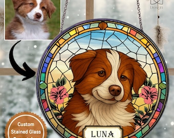 May include: A circular stained glass suncatcher featuring a brown and white dog with the name LUNA printed on a rectangular plaque at the bottom. The design includes pink flowers and a geometric border, suspended by a metal chain for display.
