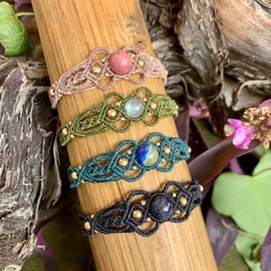 Könnte beinhalten: Vier Makramee-Armbänder mit verschiedenen farbigen Schnüren und Edelsteinperlen. Die Armbänder sind auf einer Holzoberfläche gestapelt. Die Perlen sind rosa, blau, grün und hellblau mit weißen Streifen.