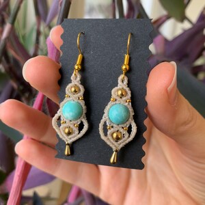 May include: A pair of turquoise gemstone earrings with gold accents and white macrame detailing. The earrings are hanging from gold hooks.