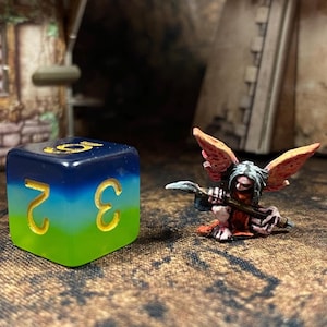 May include: A green, blue and black iridescent dice with the numbers 2 and 3 on the visible sides. A small, painted figurine of a winged creature with a red and orange body, holding a weapon, is in the background.