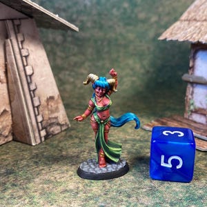 May include: A miniature fantasy figure of a female demon with blue hair and horns, wearing a green and red outfit. The figure is on a black base, with a blue die showing the number 5. The background includes miniature buildings.